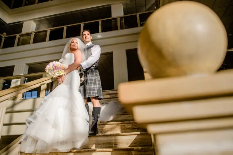 Maryellen and Ben on a curved hotel staircase beside a stone sphere, the groom in his kilt