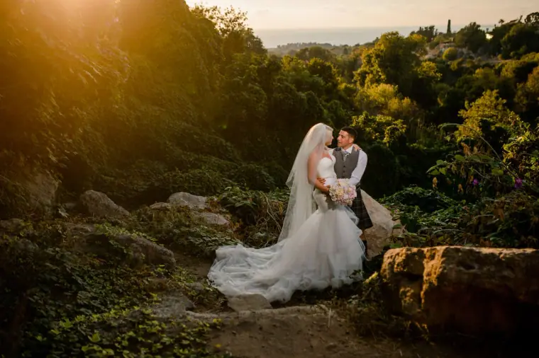 Bride and groom embracing among rocks and greenery in warm sunset light