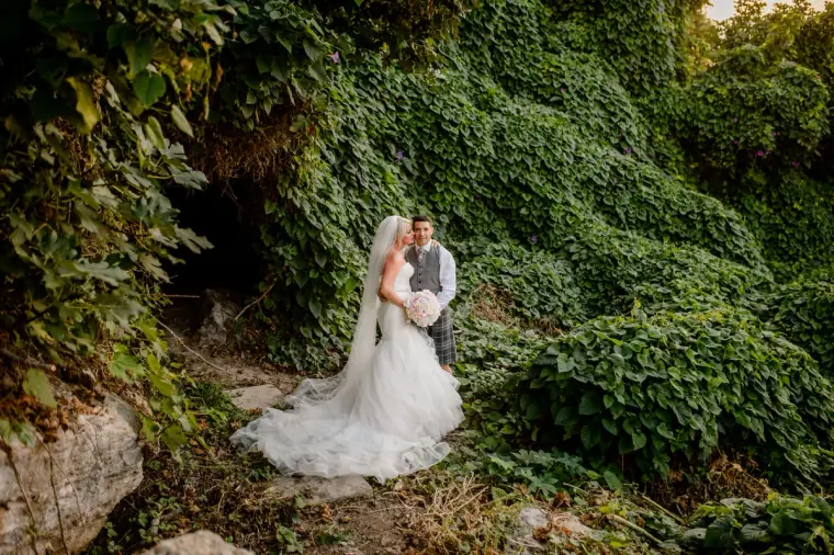 Couple embracing beside rocks and ivy, her train spread on the ground