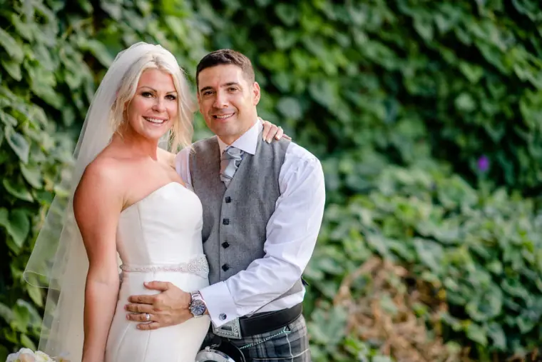 Smiling Maryellen and Ben posing against green foliage, groom in waistcoat and kilt