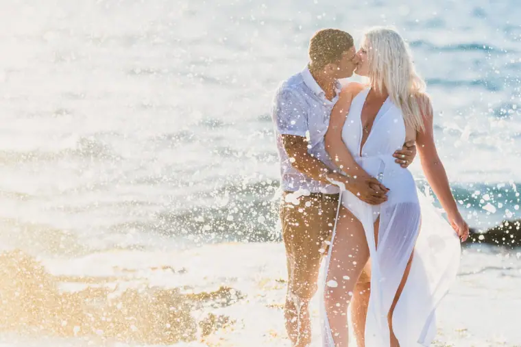 Bride in a flowing white dress kissing the groom as spray rises from the sea