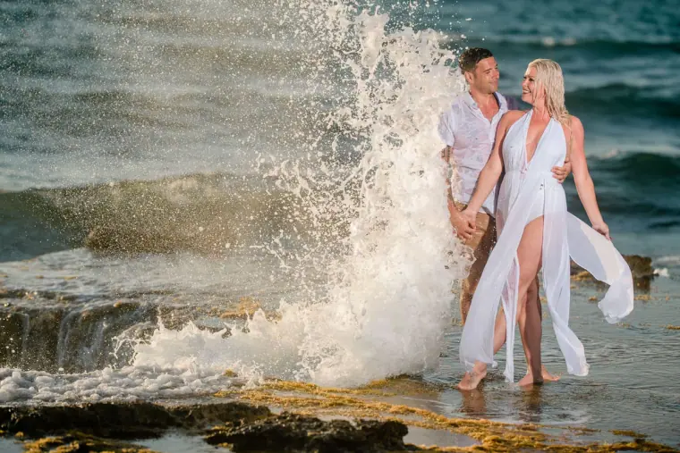 Couple laughing as a wave bursts behind them on the rocks
