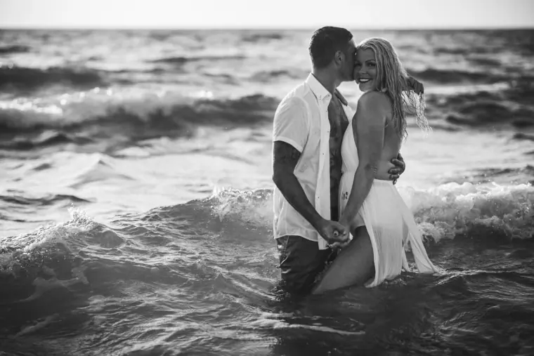 Black-and-white photo of the couple kissing in the sea as waves roll in
