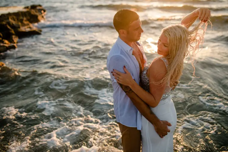 Bride lifting her wet hair as the groom holds her among the waves at sunset