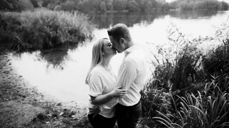 Black-and-white photo of the couple kissing among reeds by the water's edge