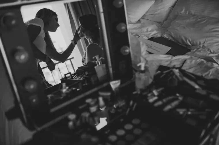Black-and-white photo of a makeup case and lit mirror reflecting the artist at work
