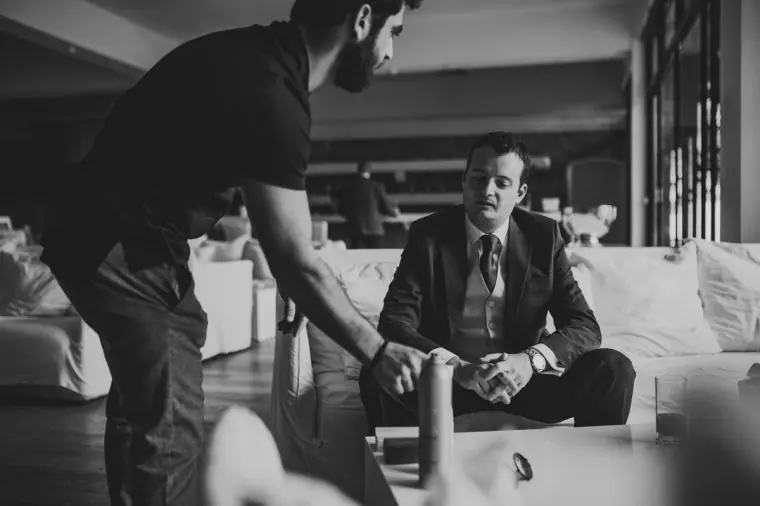 Black-and-white photo of the groom sitting on a sofa as a friend gets ready beside him