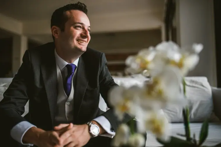 Smiling groom in a grey suit seated beside white orchids