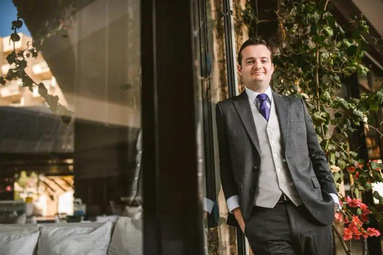 Groom in a grey three-piece suit and purple tie by a plant-lined window