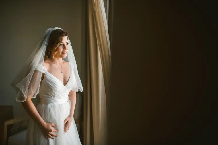 Bride gazing out of the window in her veil, light falling across her dress