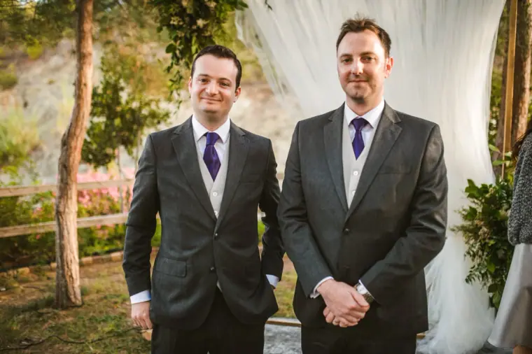 Groom and best man in grey suits and purple ties waiting by the draped ceremony arch