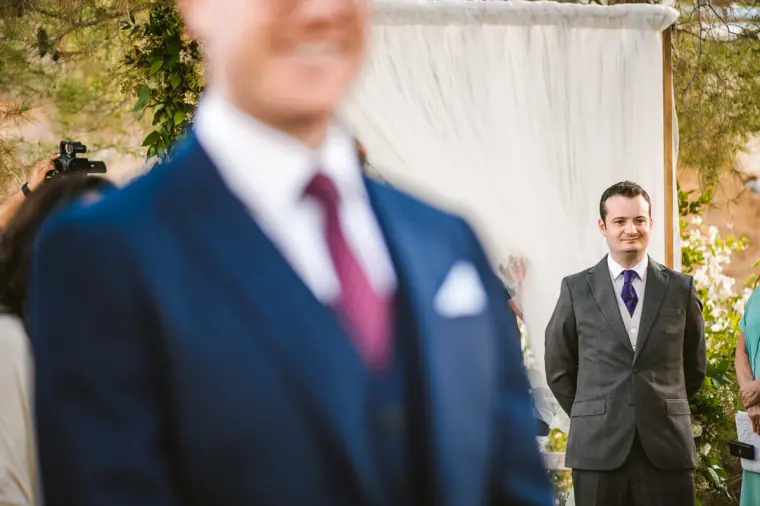 Groom waiting at the arch, seen past a blurred guest in a navy suit