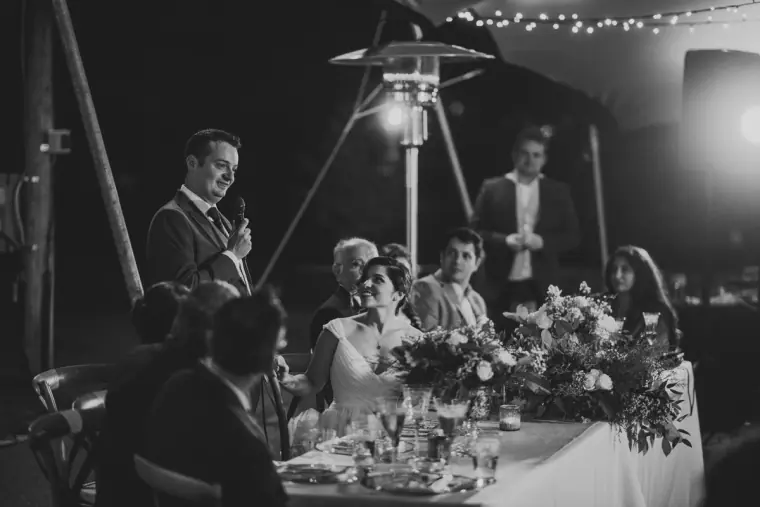 Black-and-white photo of the groom giving a speech beside the bride at the top table