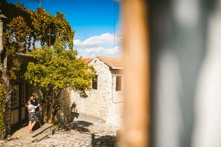 Couple glimpsed down a sunny stone lane past a blurred wooden post in Lefkara