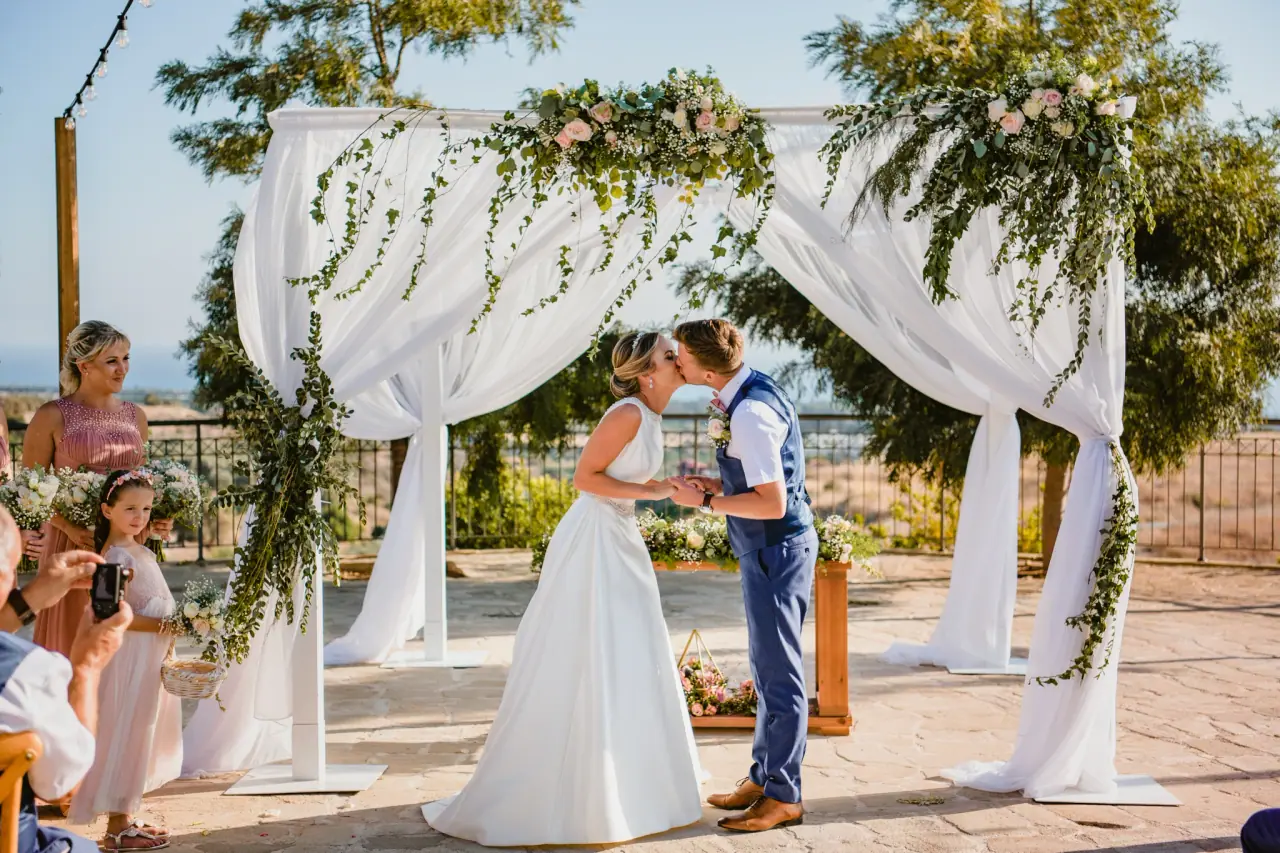Nicola and Joel kissing beneath a draped floral arch as guests look on at Liopetro, Kouklia