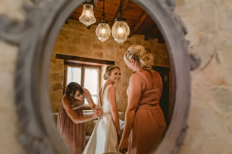 Bridesmaids fastening the bride's dress, seen in an ornate oval mirror