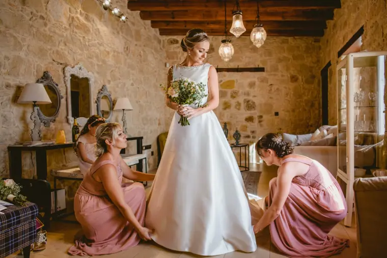 Bridesmaids in dusky pink adjusting the bride's gown in a stone-walled room at Liopetro