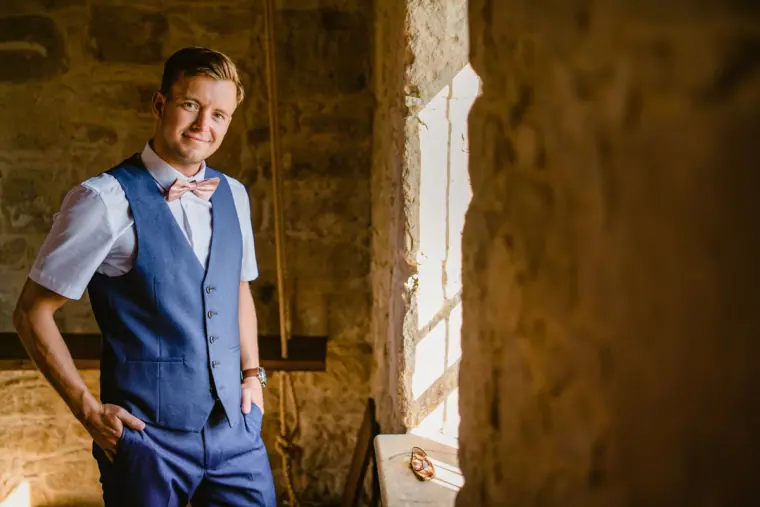 Groom in a navy waistcoat and pink bow tie standing by a sunlit window