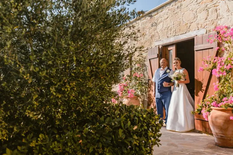 Bride and her father stepping out of a stone doorway framed by bougainvillea