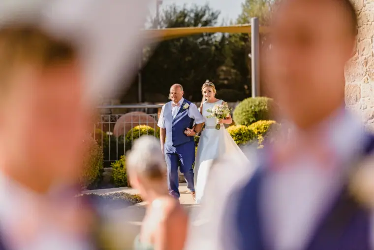 Bride walking with her father towards the ceremony between blurred guests
