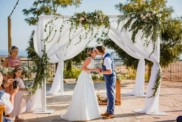 Nicola and Joel's first kiss beneath the draped arch as guests look on, Liopetro