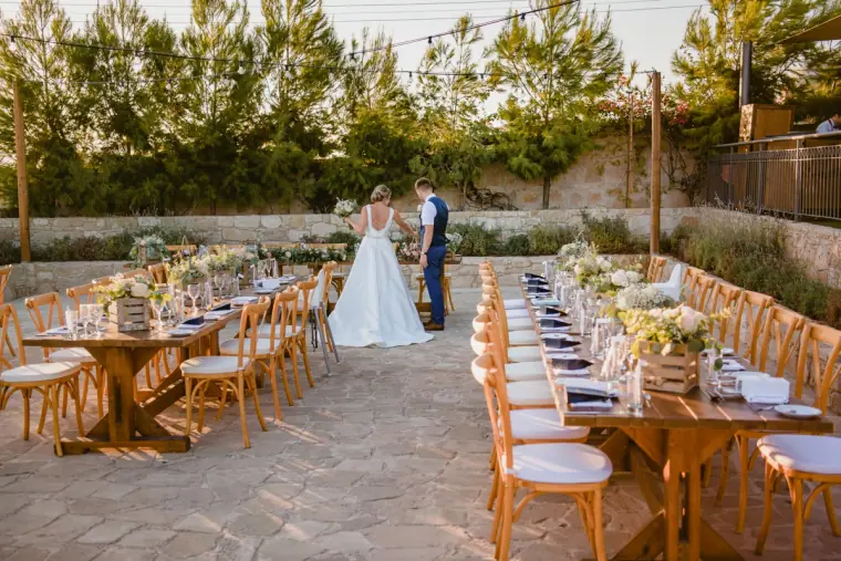 Bride and groom walking between long dining tables on the stone terrace at Liopetro