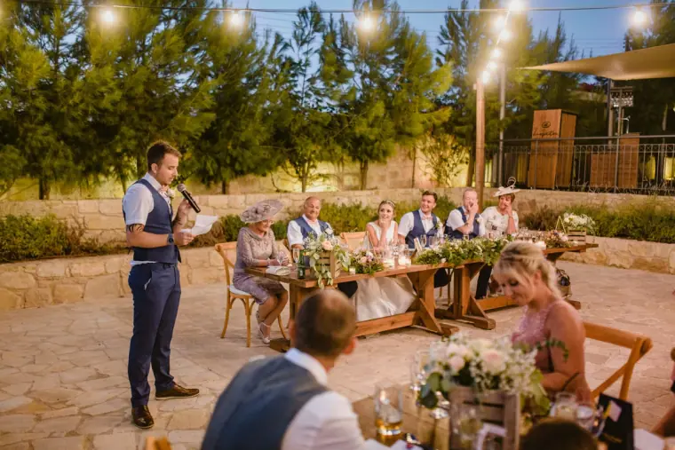 Best man giving a speech to guests at long tables under festoon lights at Liopetro, Kouklia