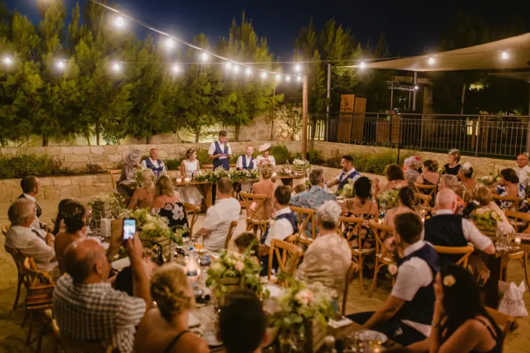 Guests at long tables listening to speeches under strings of lights at dusk