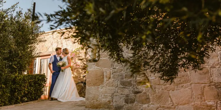 Bride and groom embracing beside a stone cottage under olive branches