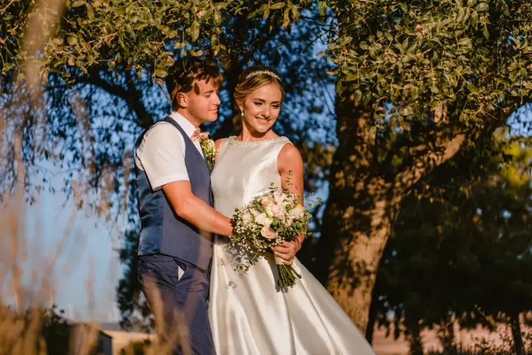 Nicola and Joel smiling under an olive tree, the bride holding her pastel bouquet