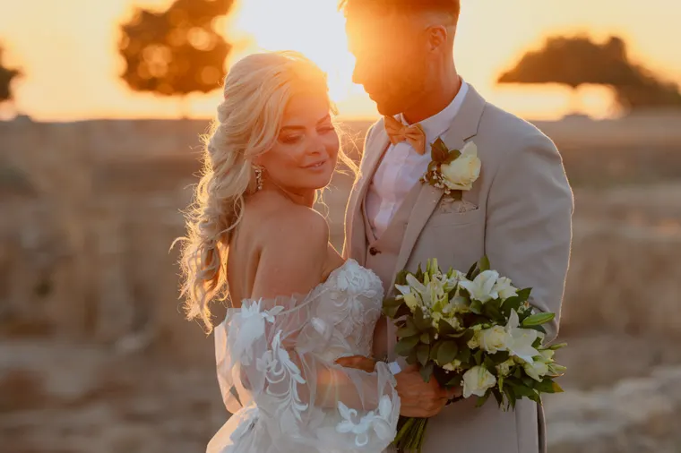 Bride and groom embracing in golden sunset light, white bouquet in hand