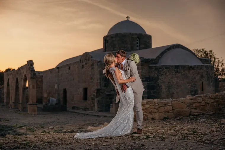 Bride and groom kissing by a domed stone church and ruins at sunset, Kouklia