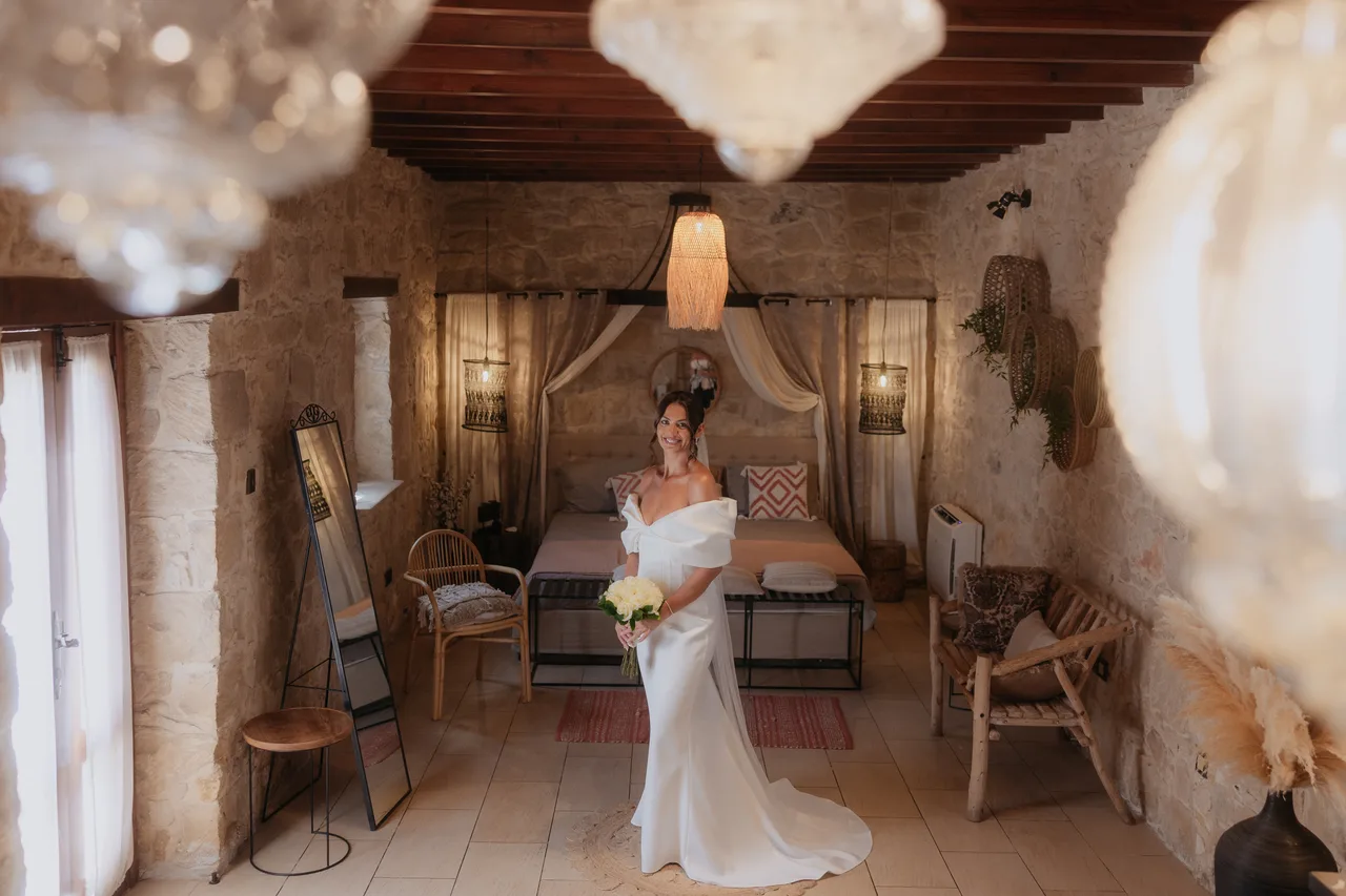 Bride in a fitted off-shoulder gown standing in a boho-styled stone suite at Liopetro