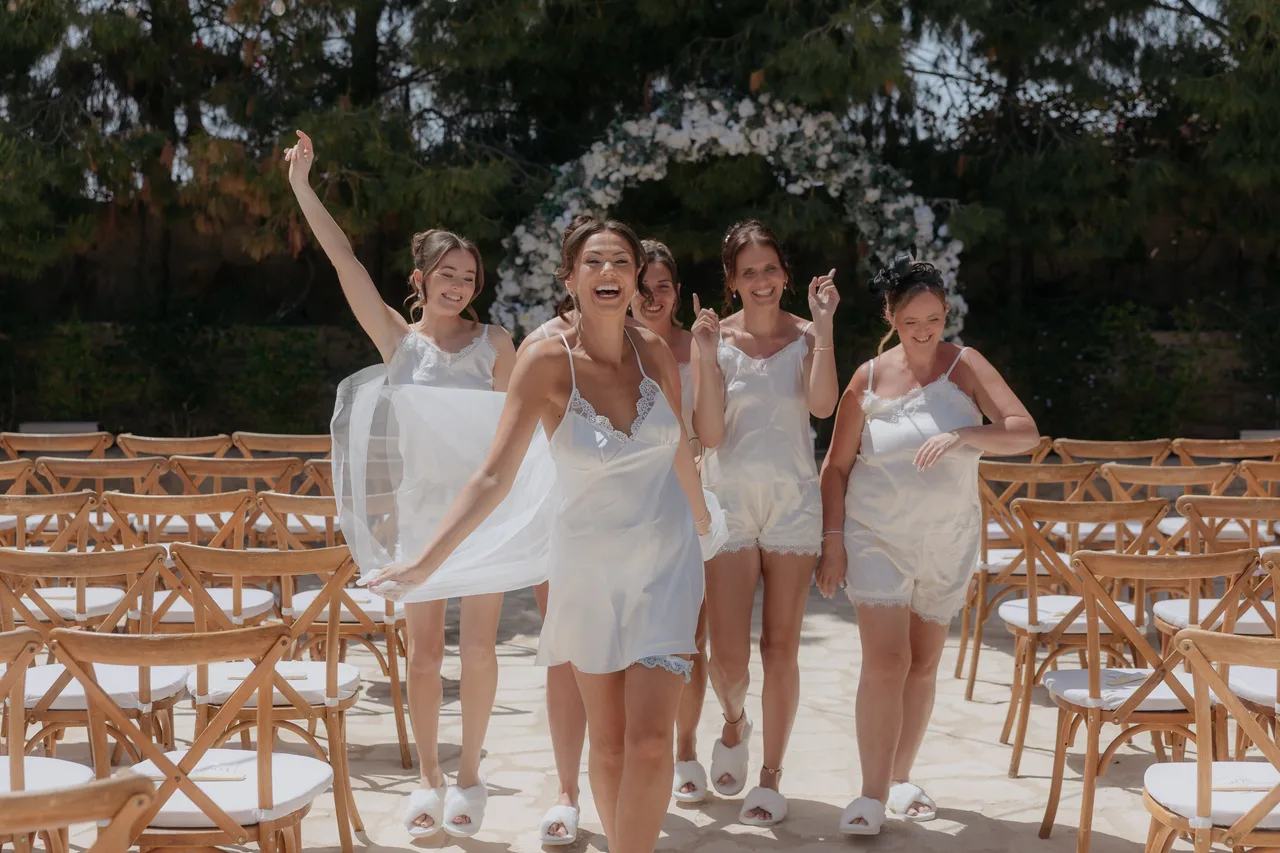 Bridesmaids in white satin cheering among ceremony chairs at Liopetro, Kouklia
