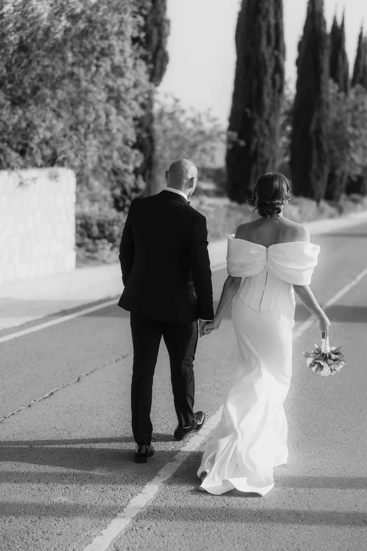 Black-and-white photo of the couple walking hand in hand along a cypress-lined road