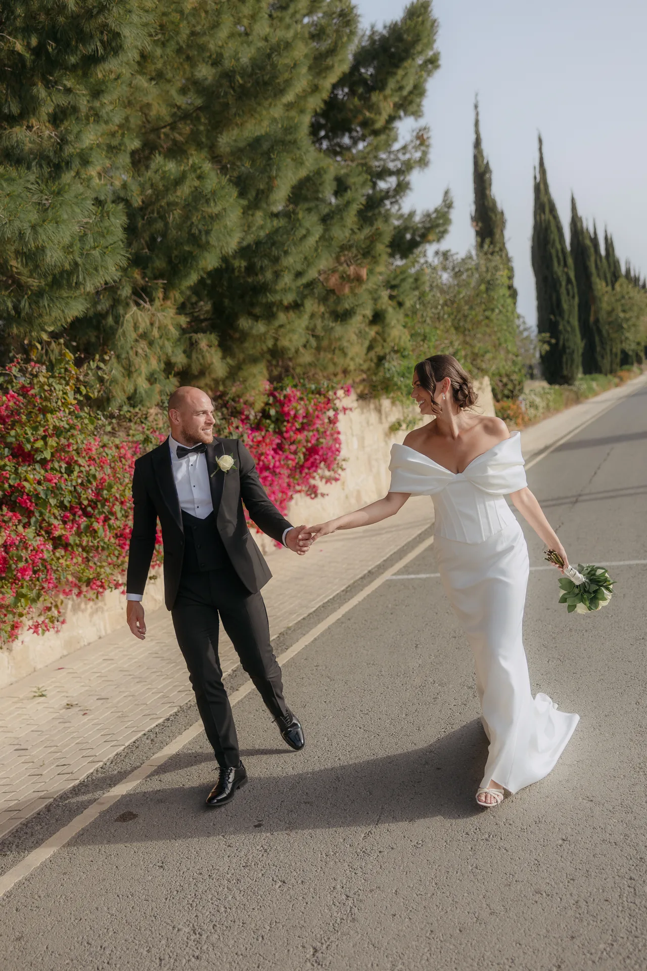 Bride and groom walking hand in hand past bougainvillea on the road near Liopetro, Kouklia
