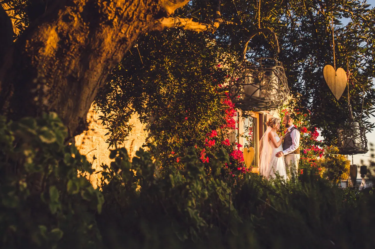 Bride and groom sharing a quiet moment through olive branches and lanterns at golden hour, Liopetro