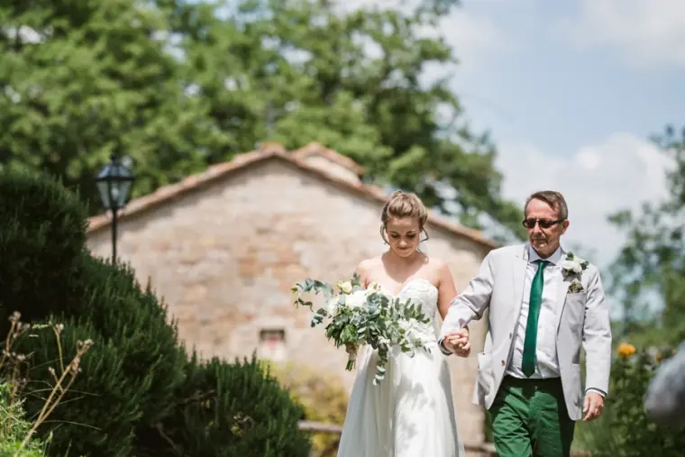 Father walking the bride to the ceremony past a stone house in the Tuscan hills
