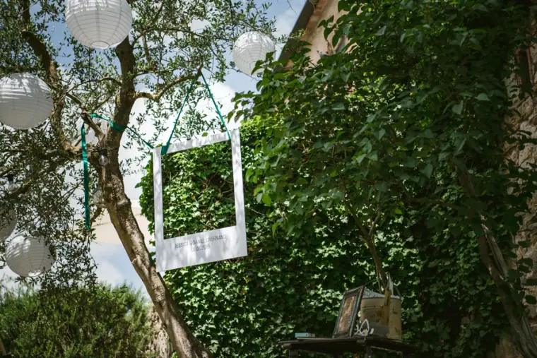 Empty white picture frame and paper lanterns hanging in trees beside an ivy-covered wall