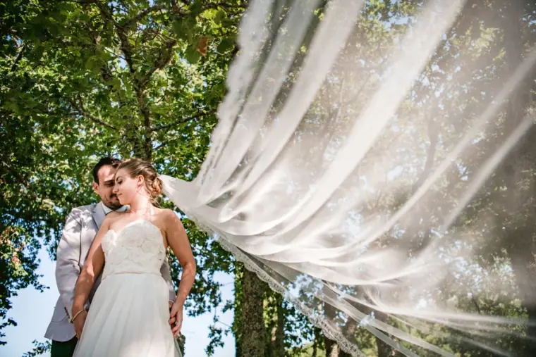 Bride's veil billowing in the wind as she stands beside her groom under trees