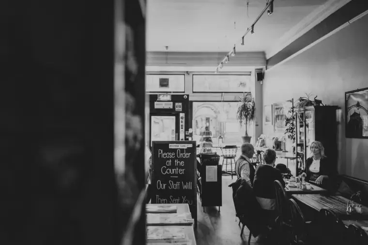 Black-and-white view of a café interior with a counter sign and customers seated by the window