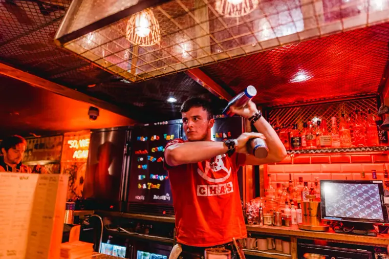 Bartender in a red T-shirt flipping a bottle behind a neon-lit bar
