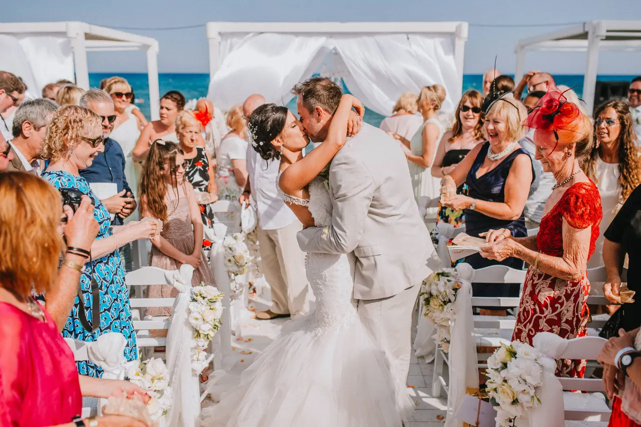 Maria and Gav kissing in the aisle as guests cheer beside the sea at Galu Seaside, Larnaca
