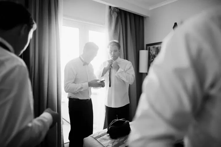 Black-and-white shot of the groom and groomsmen dressing by a bright window