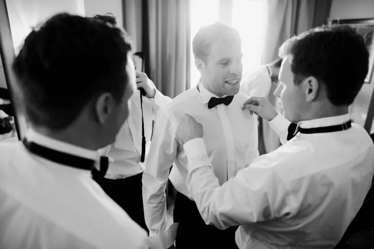 Black-and-white shot of groomsmen helping the groom with his bow tie and buttonhole