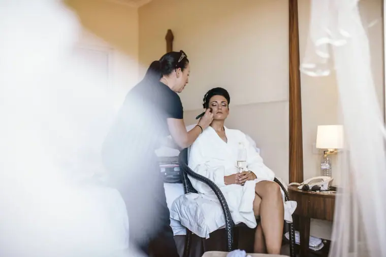 Bride in a white robe having her make-up done in a sunlit hotel room