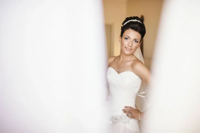 Bride in her gown and tiara framed by soft white curtains