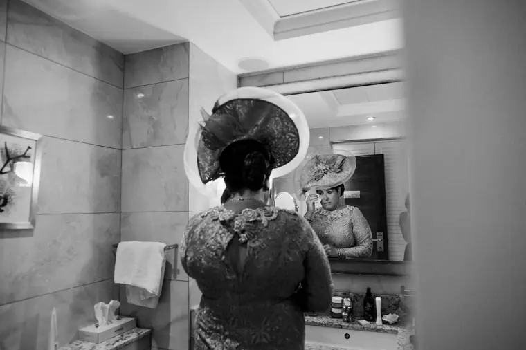 Black-and-white shot of a guest in a wide hat fixing her look in a bathroom mirror