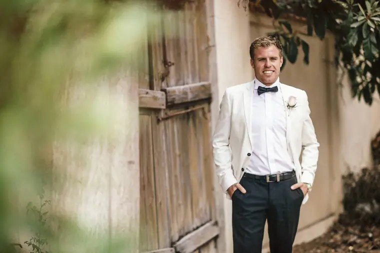 Groom in a white tuxedo jacket waiting by a weathered wooden door