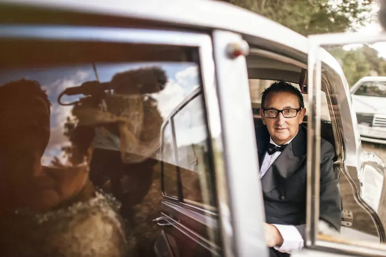 Man in a bow tie smiling through the window of the vintage wedding car